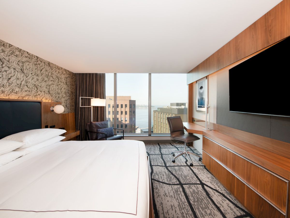 A modern hotel room with a large bed, desk, chair, big TV, and a window view of buildings and water. The decor is sleek and contemporary.