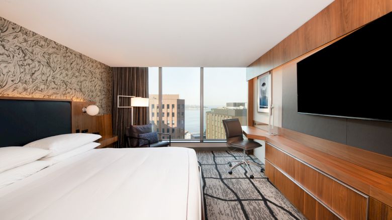 A modern hotel room with a large bed, desk, chair, big TV, and a window view of buildings and water. The decor is sleek and contemporary.