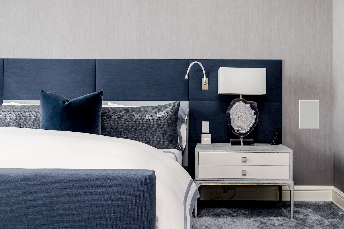 A modern bedroom with a navy and white theme features a bed, pillows, nightstand, lamp, and wall light, creating a cozy ambiance.