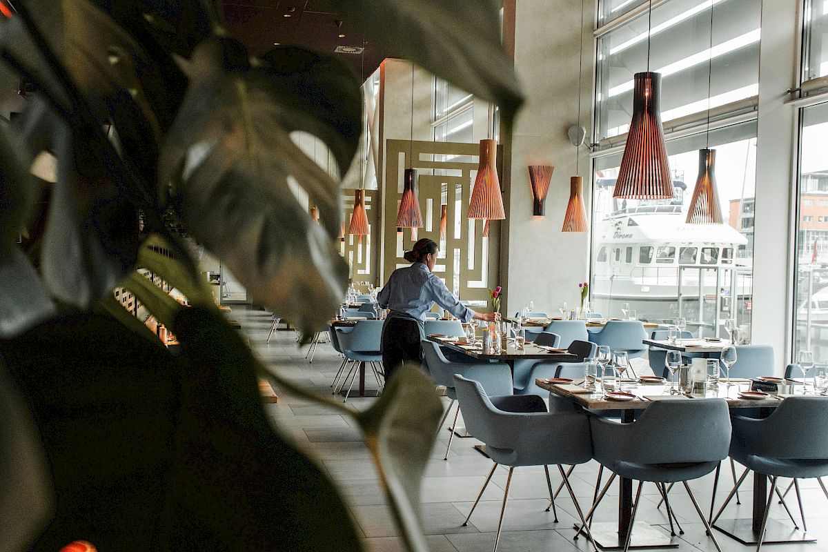 A person sets tables in a stylish, modern restaurant with large windows, pendant lights, and plants enhancing the decor.