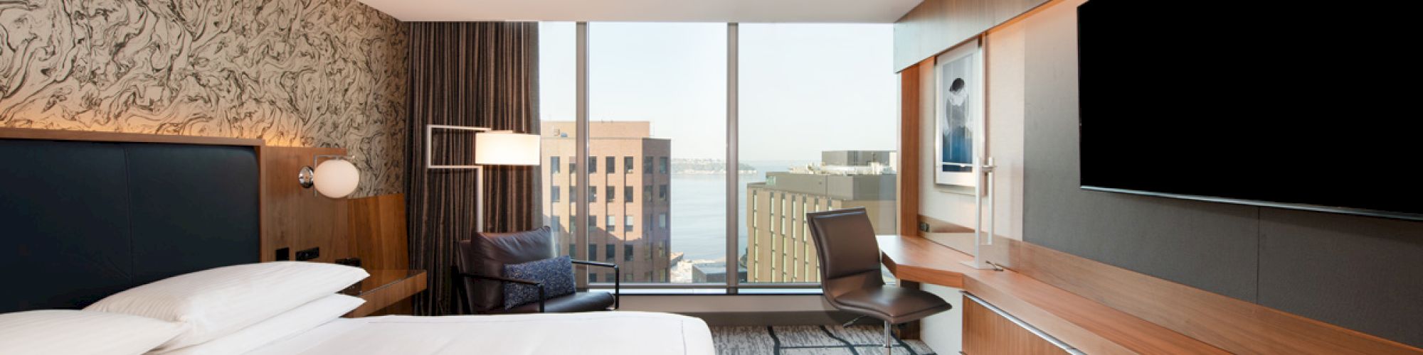 A modern hotel room features a bed, desk, chair, large TV, and window view of an urban skyline and water in the background.